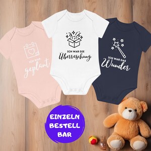 Könnte beinhalten: Drei Baby-Bodys mit unterschiedlichen Designs. Der rosa Body hat einen Kalender mit einem Herzen und den Text "Ich war geplant". Der weiße Body hat eine Geschenkbox mit Sternen und den Text "Ich war die Überraschung". Der blaue Body hat einen Zauberstab mit Sternen und den Text "Ich war das Wunder".