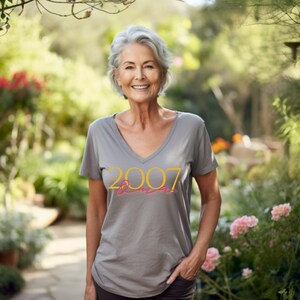 Peut inclure: Une femme portant un t-shirt gris avec le texte "Est. 2007" en jaune. Elle se tient dans un jardin avec de la v&eacute;g&eacute;tation verte et des fleurs roses.