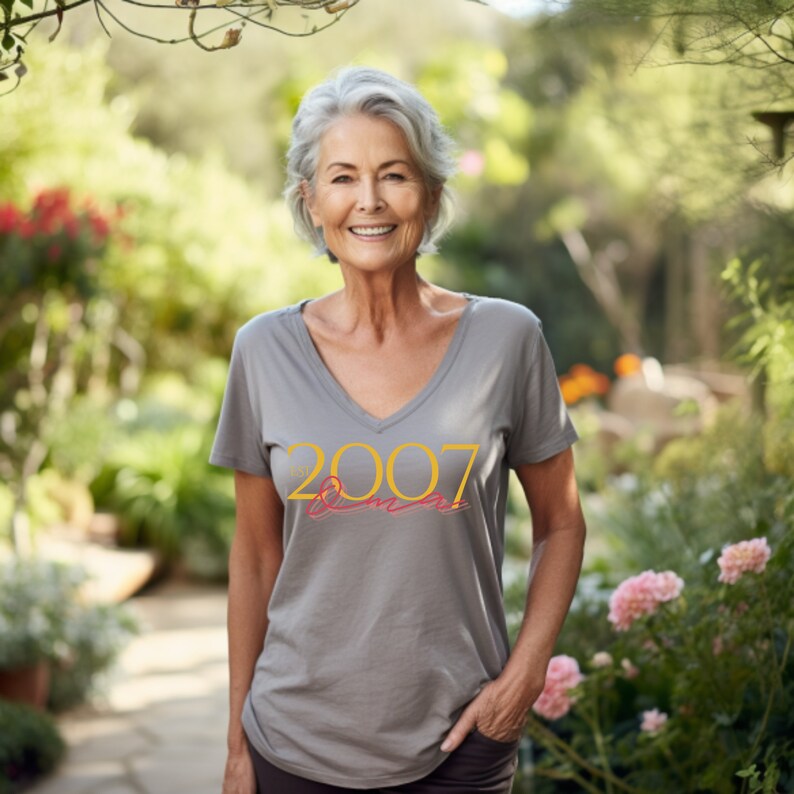 Peut inclure: Une femme portant un t-shirt gris avec le texte "Est. 2007" en jaune. Elle se tient dans un jardin avec de la v&eacute;g&eacute;tation verte et des fleurs roses.