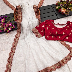 May include: White net saree with red and gold embroidered border. The saree has a scalloped edge and is paired with a red embroidered blouse.