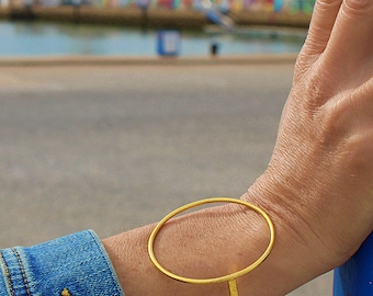 May include: A gold-colored bracelet with an oval design. The bracelet is worn on a wrist, with a denim jacket sleeve visible in the background. The bracelet's design is a large, open oval shape.
