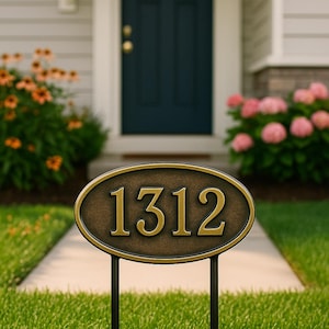 Könnte beinhalten: Ovale, bronzefarbene Hausnummerntafel mit der Nummer 1312 in goldfarbenen Buchstaben. Die Tafel ist auf zwei schwarzen Metallpfosten montiert und in einem grünen Rasen aufgestellt. Eine dunkelblaue Tür und Blumen sind im Hintergrund.