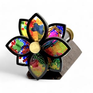 A kaleidoscopic spinning top, shaped like a flower with six petals. Each petal is filled with colorful beads, sequins, and small butterfly shapes, creating a vibrant, multicolored effect. The flower is mounted on a dark brown base. The overall design is intricate and eye-catching, suggesting a handcrafted, artistic creation. Vintage nautical brass kaleidoscope collectible. Custom engraved desk gift kaleidoscope