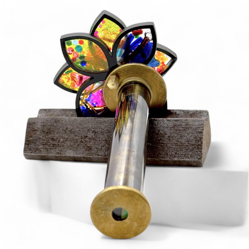 A kaleidoscope with a brass body and a colorful, flower-shaped top. The flower has petals in shades of yellow, orange, pink, blue, and green, filled with multicolored confetti-like pieces. The kaleidoscope is mounted on a dark brown wooden base.  It's a handcrafted, artistic, and visually captivating piece, perfect for decorative or playful purposes. The brass end cap has a small viewing hole. Handcrafted brass kaleidoscope closeup optics.