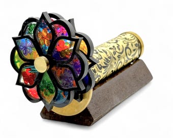 May include: A kaleidoscope with a flower-shaped end featuring colorful stained glass. The barrel is gold with a black floral pattern. It rests on a brown rectangular base. The kaleidoscope is a unique decorative item.