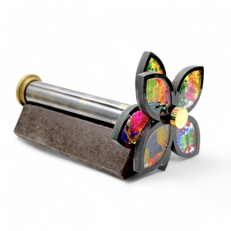 A kaleidoscopic rotating flower-shaped top on a dark brown wooden base, attached to a brushed metallic cylinder. The flower has multicolored, jewel-toned petals with embedded glitter.  The cylinder is dark gray with a brass-colored end cap. The overall design is compact and decorative, suitable as a unique home decor item or a mesmerizing toy. Engraving name and date on brass kaleidoscope body