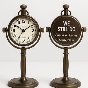 May include: Two bronze-colored table clocks. One clock face shows the time, the other has the text "WE STILL DO" with names and a date. The clocks have a round base and a decorative top.