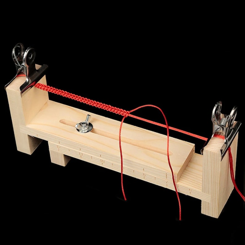 Op de afbeelding: Een houten armbandweefgetouw met een rood koord erdoorheen geregen. Het weefgetouw heeft twee klemmen en een schuifbalk met een knop. Het koord is om het weefgetouw gewikkeld en vastgezet met de klemmen.