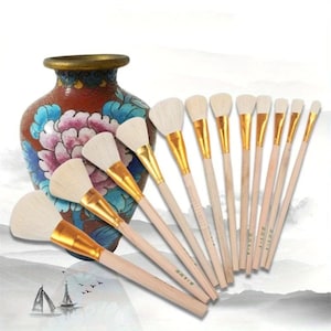 May include: A set of 12 fan-shaped paint brushes with white bristles and gold-colored ferrules. The brushes have wooden handles and are arranged in a fan shape. The brushes are displayed in front of a decorative vase with floral designs.