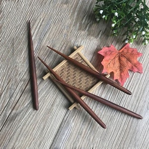 May include: Three dark brown wooden hair sticks on a woven bamboo mat with a red maple leaf and green foliage in the background.
