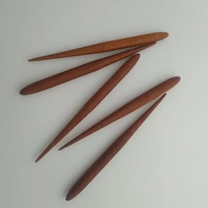 May include: Five wooden knitting needles, all with pointed tips and a natural wood finish. The needles are arranged in a fan shape, with the tips pointing towards the top of the image.