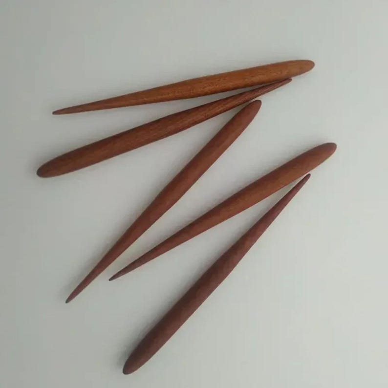 May include: Five wooden knitting needles, all with pointed tips and a natural wood finish. The needles are arranged in a fan shape, with the tips pointing towards the top of the image.