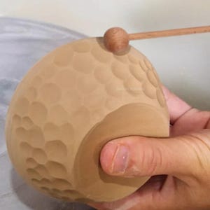 Op de afbeelding: Een bruine keramische kom met een getextureerd oppervlak. De kom wordt vastgehouden door een hand en een houten gereedschap wordt gebruikt om de textuur te creëren.