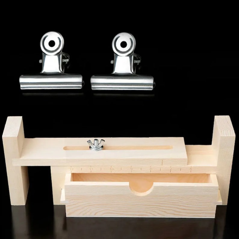 Op de afbeelding: Een houten zeepvorm met een ingebouwde lade en twee metalen klemmen. De vorm heeft een liniaal aan de bovenkant en een kleine knop om de hoogte van de vorm aan te passen.