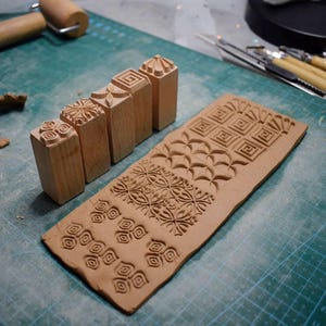 May include: A selection of wooden clay stamps featuring diverse geometric and floral designs. The stamps are positioned beside a clay slab showcasing the imprinted patterns. The stamps are light brown, and the clay is reddish-brown.