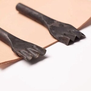 Pu&ograve; includere: Due strumenti in metallo per la lavorazione del cuoio con bordi di taglio di forme diverse. Gli strumenti sono appoggiati su un pezzo di cuoio marrone chiaro.