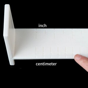 Op de afbeelding: Witte kunststof liniaal met inch- en centimetermarkeringen. De liniaal is L-vormig en heeft een vlak oppervlak.