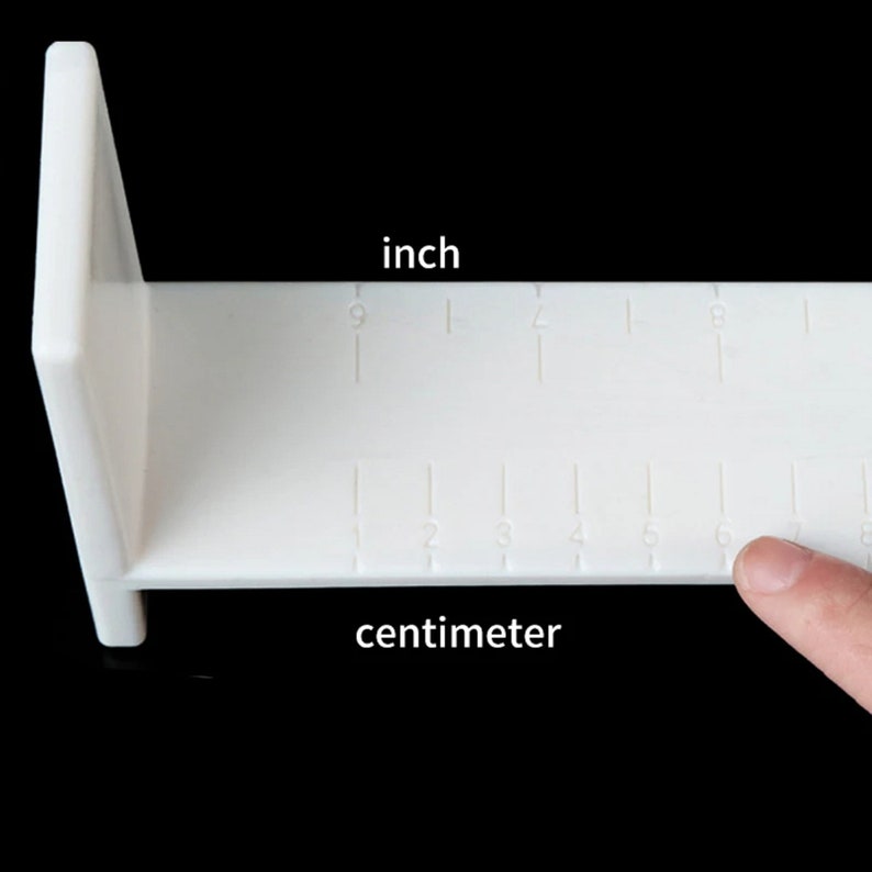 Op de afbeelding: Witte kunststof liniaal met inch- en centimetermarkeringen. De liniaal is L-vormig en heeft een vlak oppervlak.