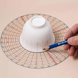 May include: A white ceramic bowl is placed on a circular gridded template with red and black lines. A blue pencil is used to draw lines on the bowl, following the grid lines on the template.