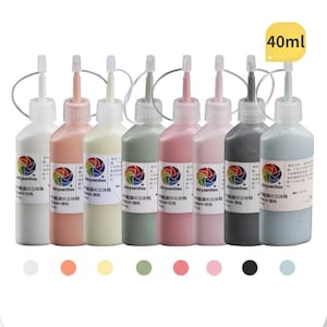 Peut inclure: Huit flacons souples remplis de peinture aux couleurs pastel variées. Les flacons, chacun d'une teinte différente, sont disposés en rangée. Les couleurs comprennent le blanc, le pêche et le gris. Chaque flacon porte une étiquette avec le mot "Chrysanthos" et un logo multicolore. Une étiquette jaune indique une capacité de 40 ml.