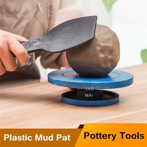 Puede incluir: Una persona usa una herramienta de plástico negro para dar forma a una bola de arcilla marrón sobre un torno de alfarero azul. La rueda está sobre una superficie de madera. El texto "Plastic Mud Pat Pottery Tools" está en la parte inferior.