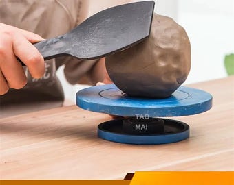 May include: A person using a black plastic tool to shape a ball of brown clay on a blue pottery wheel. The wheel is on a wooden surface. The text "Plastic Mud Pat Pottery Tools" is at the bottom.