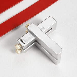 May include: A silver leather cutting tool with a ruler and two adjustable knobs. The tool is used to cut leather strips to a specific width.
