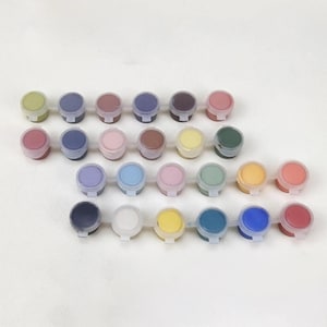 Puede incluir: Un conjunto de 24 pequeños botes de pintura con tapas, dispuestos en filas. Los colores de la pintura incluyen tonos de verde, azul, morado, rosa, amarillo, marrón, rojo y blanco.