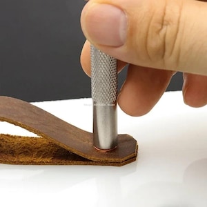 May include: A metal leather punch tool with a textured handle is being used to create a hole in a piece of brown leather. The tool is silver and the leather is a warm brown color. The image is a close-up.