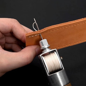 May include: A close-up of a leather sewing awl with a white thread spool and a needle threading through a piece of brown leather.