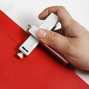 Könnte beinhalten: Ein silbernes Werkzeug aus Metall mit einem Lineal und einer Klinge zum Schneiden von Leder. Das Werkzeug wird verwendet, um ein Stück rotes Leder zu schneiden.