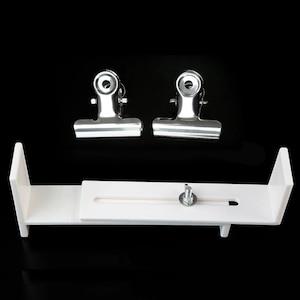 Op de afbeelding: Een witte plastic achtergrondstandaard met een schuifbalk en een schroef. Twee zilveren metalen clips zijn inbegrepen.