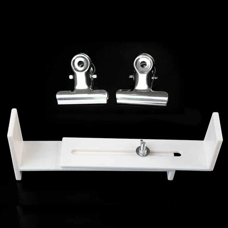 Op de afbeelding: Een witte plastic achtergrondstandaard met een schuifbalk en een schroef. Twee zilveren metalen clips zijn inbegrepen.