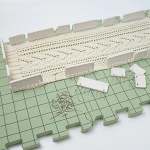 Può includere: Ferri da maglia bianchi e un telaio da maglia bianco su una superficie verde a griglia. Un pezzo all'uncinetto è sul telaio. Ci sono anche ferri da maglia argentati sulla superficie.