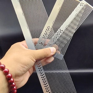 May include: Four metal stencils with a grid of small holes are displayed. Each stencil is rectangular with rounded corners and the number "139" printed on it. The stencils are held in a hand, and a red beaded bracelet is visible.