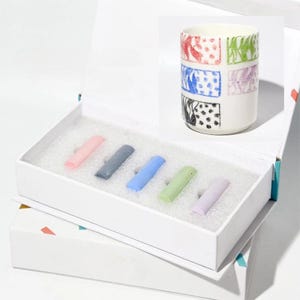 May include: A white gift box with a set of six pastel-colored sticks and a matching ceramic cup. The cup features a colorful, geometric design with red, green, blue, and black patterns. The box is open, revealing the contents.