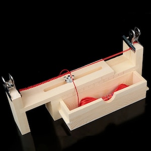 Op de afbeelding: Een houten gereedschap voor het maken van armbanden met een rood koord dat erdoorheen is geregen. Het gereedschap heeft een ingebouwde liniaal en een klein lade om het koord op te bergen.