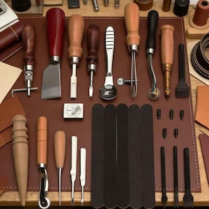 Könnte beinhalten: Eine Sammlung von Lederwerkzeugen, darunter Messer, Ahlen, Kantenformer und ein Nahtriller, angeordnet auf einer braunen Lederoberfläche. Die Werkzeuge haben Holzgriffe und Metallklingen in verschiedenen Formen und Größen.