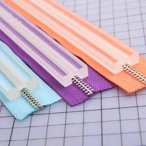 May include: Close-up of three colorful zippers: light blue, purple, and orange. Each zipper has a clear plastic slider and silver metal teeth. The zippers are arranged diagonally against a grid background, showcasing sewing supplies.