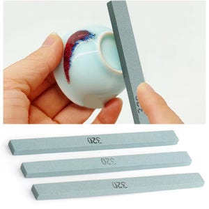 Puede incluir: Una taza de cerámica azul claro con detalles en rojo y azul se está afilando con una piedra de afilar gris. También se muestran tres piedras de afilar grises adicionales con el número "320" impreso.