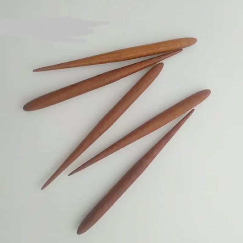 May include: Five wooden knitting needles of varying lengths, with pointed tips and rounded ends. The needles are arranged in a criss-cross pattern on a white surface.