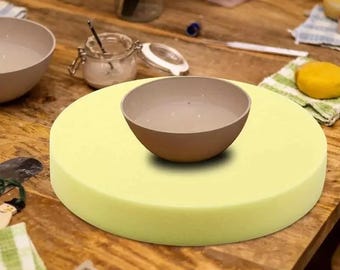 May include: A light yellow round foam turntable for pottery, with a light brown ceramic bowl on top.
