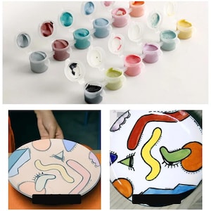 May include: A set of 18 small paint pots with lids, arranged in a row. The pots contain various colors of paint, including red, blue, green, yellow, and black. The paint pots are on a white surface. Two plates are shown, one with a partially painted design and the other with a completed design. The completed design features a colorful abstract pattern with geometric shapes and lines.