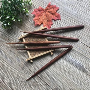 May include: Five wooden hair sticks with pointed ends. The sticks are a dark reddish-brown color and are arranged on a small wooden stand.