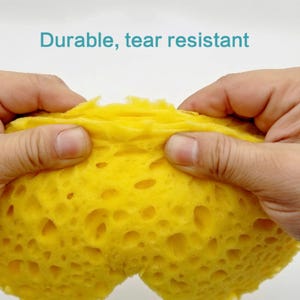 May include: Close-up of a yellow sponge being stretched and pulled apart to show its durability and tear resistance. The sponge has a honeycomb-like texture.