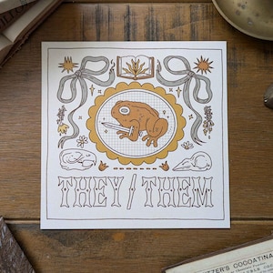 May include: A white card with a brown frog holding a sword in a yellow and brown frame. The card has a decorative border with bows and flowers. The text "They/Them" is written in brown letters at the bottom of the card.