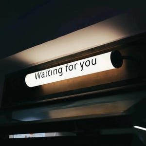 May include: A rectangular, illuminated sign with the words "Waiting for you" in black lettering. The sign is white and mounted on a dark wooden surface. The background is dark, with a soft glow emanating from the sign.