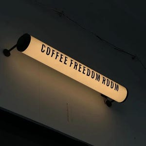 May include: A cylindrical, illuminated sign displaying the words "COFFEE FREEDOM ROOM" in black text. The sign is mounted on a wall with black fixtures. The sign is lit internally, emitting a warm light.