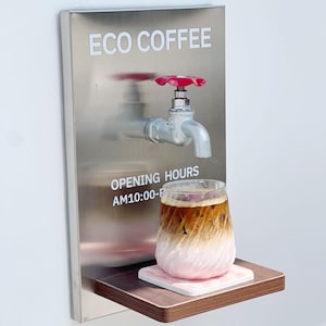 Puede incluir: Un accesorio de pared de acero inoxidable con un grifo y un mango rojo, etiquetado como "ECO COFFEE" y "OPENING HOURS AM10:00-F". Un vaso de café en capas se encuentra en un estante de madera con un posavasos rosa.