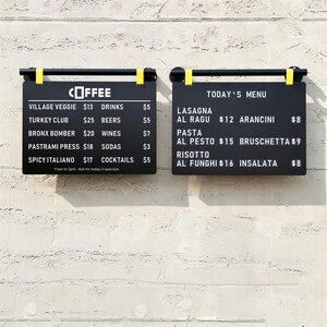 May include: Two rectangular black menu boards with white text mounted on a wall. The left board lists coffee and food items like Village Veggie and Turkey Club. The right board displays Today's Menu with items including Lasagna and Risotto.